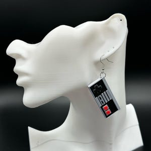 Puede incluir: Pendientes plateados con un diseño de mando negro y gris y detalles en rojo. Los pendientes tienen la forma de un mando de videojuego clásico, con un botón en forma de cruz y botones rectangulares.