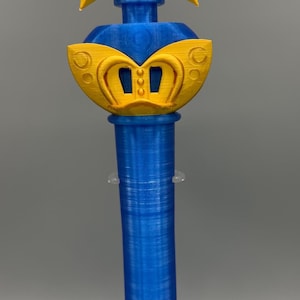 K&ouml;nnte beinhalten: Ein 3D-gedruckter Zauberstab mit einem leuchtend gelben Stern an der Spitze und einem zentralen blauen kreisf&ouml;rmigen Emblem, das das Saturnsymbol zeigt. Der Griff ist ein zylindrischer blauer Schaft mit einem gelben fl&uuml;gelartigen Element.