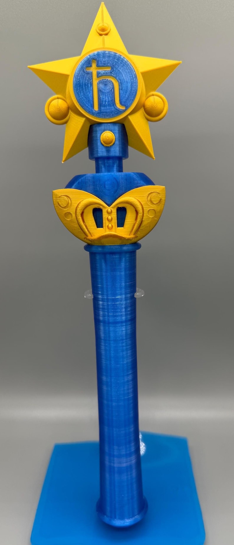 K&ouml;nnte beinhalten: Ein 3D-gedruckter Zauberstab mit einem leuchtend gelben Stern an der Spitze und einem zentralen blauen kreisf&ouml;rmigen Emblem, das das Saturnsymbol zeigt. Der Griff ist ein zylindrischer blauer Schaft mit einem gelben fl&uuml;gelartigen Element.