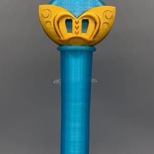 K&ouml;nnte beinhalten: Ein 3D-gedruckter Zauberstab mit einem leuchtend gelben Stern an der Spitze, der ein hellblaues kreisf&ouml;rmiges Emblem mit einem Planetensymbol enth&auml;lt. Der Griff ist ein zylindrischer hellblauer Schaft und die Basis hat ein gelbes, kronenartiges Element.