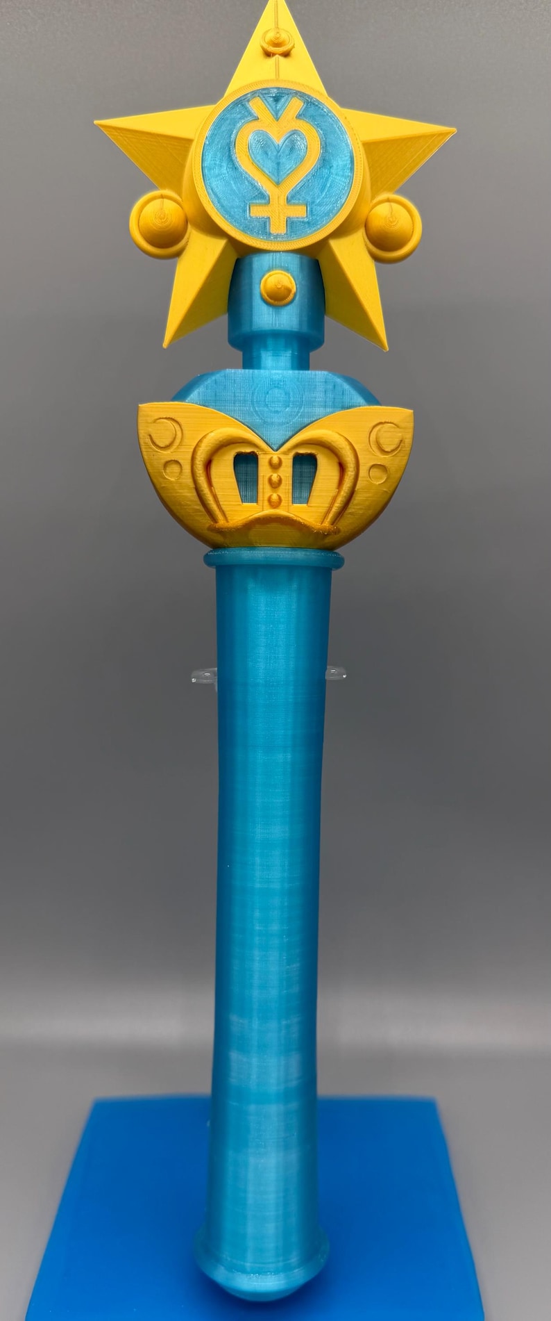 K&ouml;nnte beinhalten: Ein 3D-gedruckter Zauberstab mit einem leuchtend gelben Stern an der Spitze, der ein hellblaues kreisf&ouml;rmiges Emblem mit einem Planetensymbol enth&auml;lt. Der Griff ist ein zylindrischer hellblauer Schaft und die Basis hat ein gelbes, kronenartiges Element.
