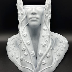 May include: A light gray decorative vase in the shape of a face with hands covering the ears. The hands and face are adorned with a vine and leaf pattern. The vase is made of a textured material.