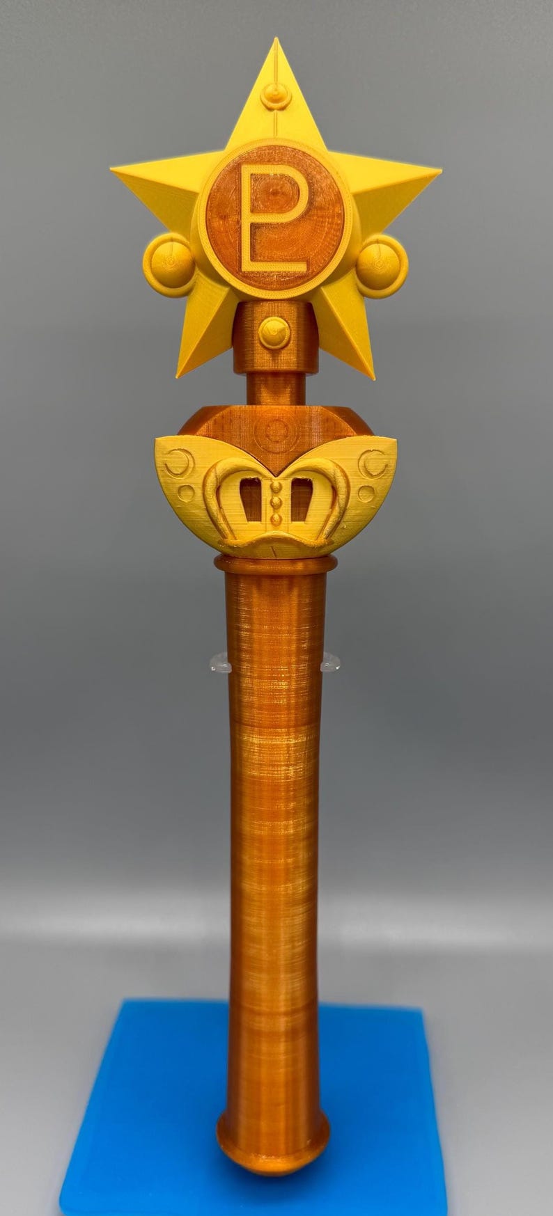 K&ouml;nnte beinhalten: Ein 3D-gedrucktes Zepter mit einem metallisch orangefarbenen zylindrischen Griff und einem gelben sternf&ouml;rmigen Aufsatz. In der Mitte des Sterns befindet sich ein kreisf&ouml;rmiges Emblem mit dem Buchstaben P, erg&auml;nzt durch eine geschwungene gelbe Basis.