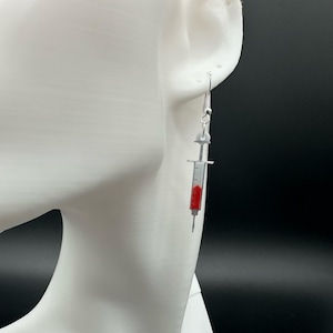 May include: Silver syringe earrings with red liquid inside. The earrings are hanging from a white ear model against a black background. The syringe design includes a needle and measurement markings.