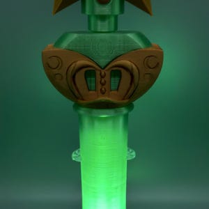 K&ouml;nnte beinhalten: Ein dekorativer Zauberstab mit einem goldenen Stern an der Spitze und einem zentralen gr&uuml;nen runden Emblem, das das Jupiter-Planetensymbol zeigt. Der Griff ist ein durchscheinender, leuchtend gr&uuml;ner Zylinder mit goldenen Akzenten am Sockel.