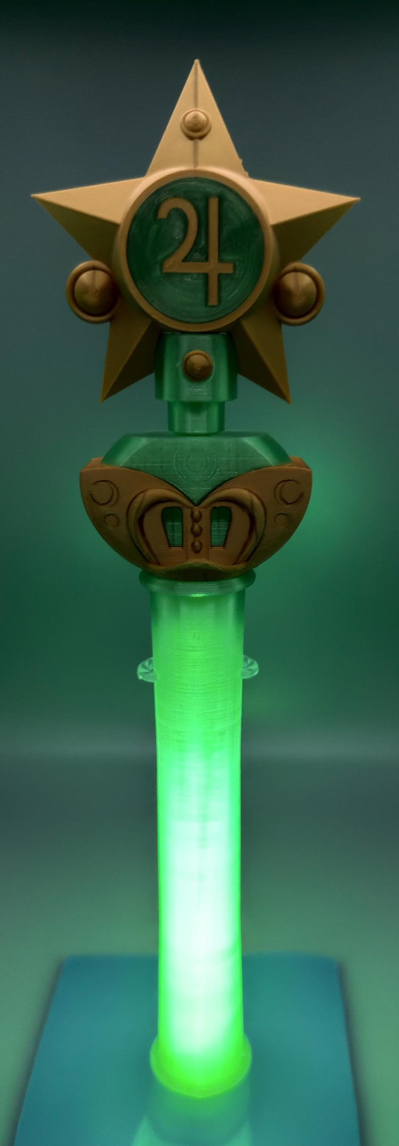 K&ouml;nnte beinhalten: Ein dekorativer Zauberstab mit einem goldenen Stern an der Spitze und einem zentralen gr&uuml;nen runden Emblem, das das Jupiter-Planetensymbol zeigt. Der Griff ist ein durchscheinender, leuchtend gr&uuml;ner Zylinder mit goldenen Akzenten am Sockel.