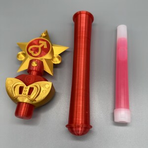 K&ouml;nnte beinhalten: Ein dreiteiliges Set bestehend aus einem gold-roten sternf&ouml;rmigen Zauberstabaufsatz mit Herz- und Pfeilsymbol, einem langen roten zylindrischen Griff und einem schlanken, durchscheinenden rosa R&ouml;hrchen mit wei&szlig;er Kappe, alle mit 3D-Druck-Struktur.