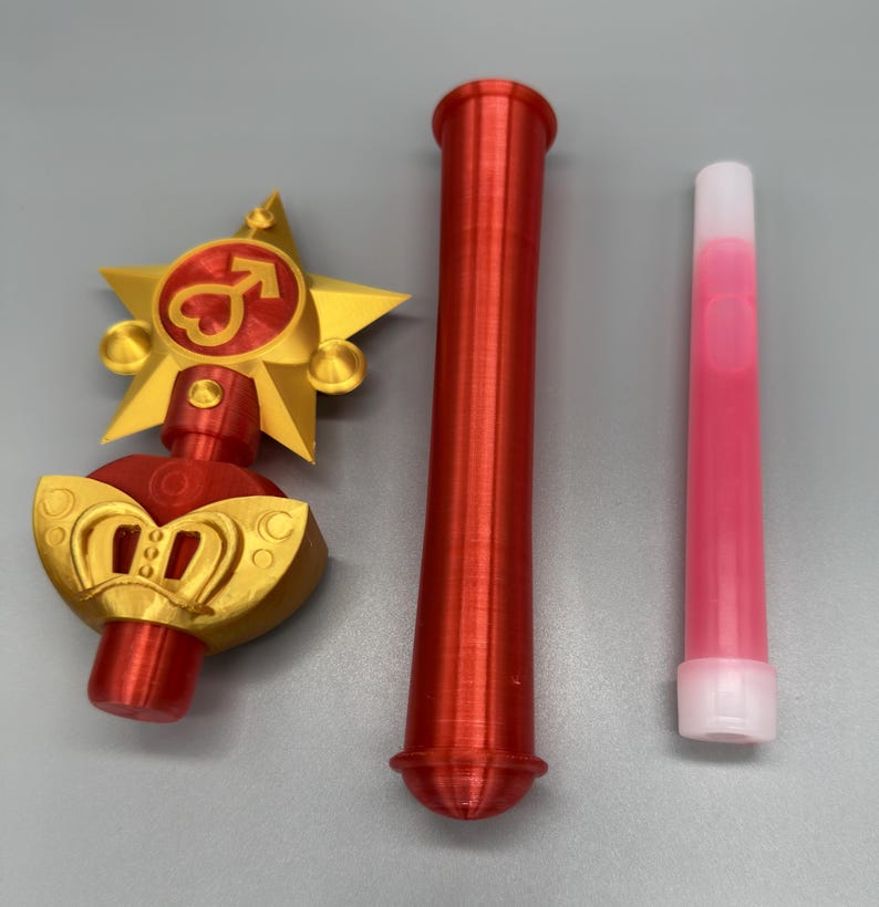 K&ouml;nnte beinhalten: Ein dreiteiliges Set bestehend aus einem gold-roten sternf&ouml;rmigen Zauberstabaufsatz mit Herz- und Pfeilsymbol, einem langen roten zylindrischen Griff und einem schlanken, durchscheinenden rosa R&ouml;hrchen mit wei&szlig;er Kappe, alle mit 3D-Druck-Struktur.