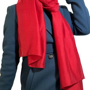 May include: A bright red silk scarf draped over a teal blue coat. The scarf is folded in half and worn around the neck.