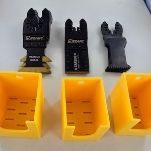 Könnte beinhalten: Drei gelbe, wandmontierte Aufbewahrungshalter aus Kunststoff mit schwarzen oszillierenden Werkzeugklingen. Die Klingen sind mit "Titanium Metal", "Carbide" und "CEZARC" beschriftet.