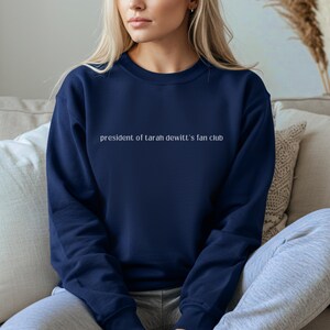 May include: A navy blue crew neck sweatshirt featuring the white text "president of tarah dewitt's fan club". The sweatshirt is made of a soft material.