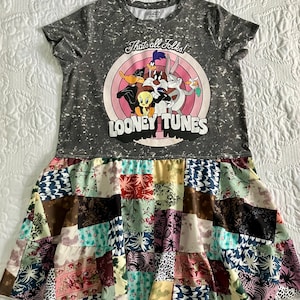 May include: A grey Looney Tunes dress with a patchwork skirt in various colours. The top has a speckled design and a circular graphic featuring Looney Tunes characters and the words "That's all Folks!".