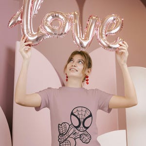May include: A mauve t-shirt featuring a black graphic of Spider-Man. Above, a person holds a pink metallic balloon spelling "LOVE". The background is a pink, abstract design.