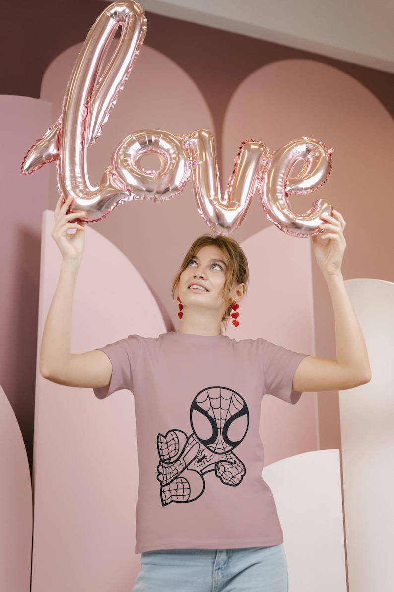 May include: A mauve t-shirt featuring a black graphic of Spider-Man. Above, a person holds a pink metallic balloon spelling "LOVE". The background is a pink, abstract design.