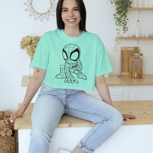 May include: A light teal crop top featuring a black and white cartoon Spider-Man graphic. The shirt is paired with light wash denim jeans. The overall aesthetic is casual and youthful.