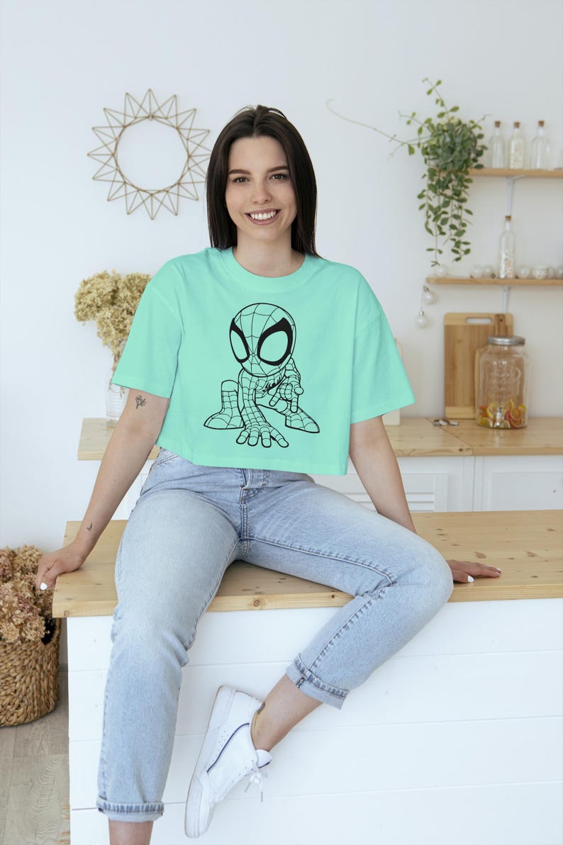 May include: A light teal crop top featuring a black and white cartoon Spider-Man graphic. The shirt is paired with light wash denim jeans. The overall aesthetic is casual and youthful.