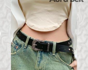 May include: A black leather belt with a silver buckle featuring an ornate design. The belt has small silver studs along the edge. The belt is worn over a pair of light green denim jeans.