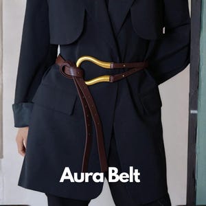 May include: A brown leather belt with a gold-colored buckle is wrapped around a dark navy blue coat. The belt has a unique design with a curved buckle and a long, dangling strap. The text "Aura Belt" is displayed in white.