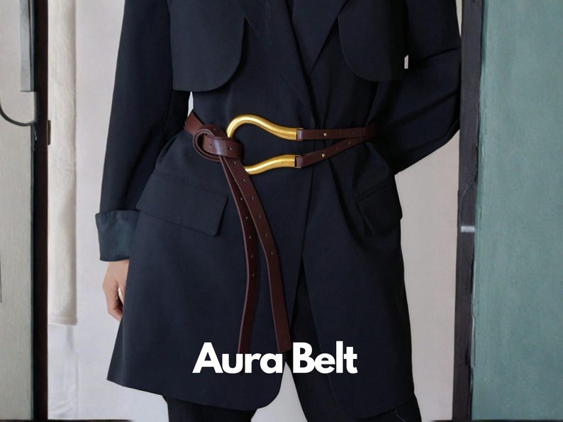 May include: A brown leather belt with a gold-colored buckle is wrapped around a dark navy blue coat. The belt has a unique design with a curved buckle and a long, dangling strap. The text "Aura Belt" is displayed in white.