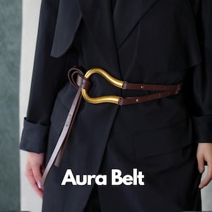 Puede incluir: Un cinturón de cuero marrón con una hebilla dorada de diseño curvo único. El cinturón se lleva sobre un abrigo negro. El texto "Aura Belt" se muestra en blanco en la parte inferior de la imagen.