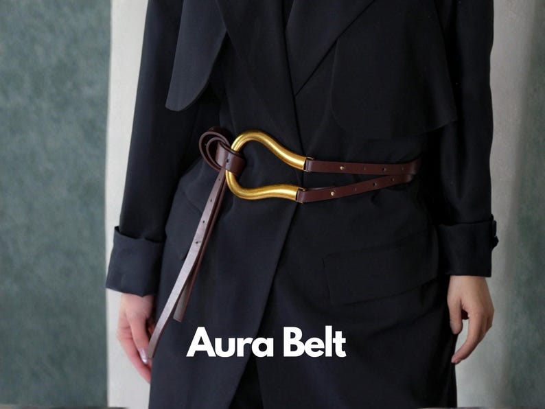 May include: A brown leather belt with a gold-colored buckle in a unique, curved design. The belt is worn over a black coat. The text "Aura Belt" is displayed in white at the bottom of the image.