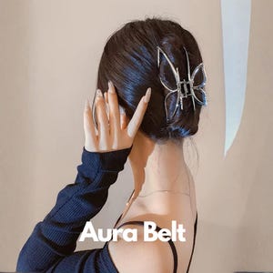 Puede incluir: Un clip de pelo plateado con forma de mariposa se lleva en el pelo. El texto "Aura Belt" es visible en la parte inferior de la imagen. La persona lleva una camiseta negra de manga larga.