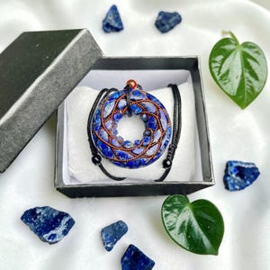 May include: A circular pendant featuring blue lapis lazuli stones wrapped in copper wire, suspended on a black cord. The pendant rests on a white cushion inside a black gift box, accompanied by loose blue stone fragments and green leaves.