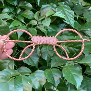 May include: A pair of decorative glasses crafted from twisted copper wire, featuring a central horizontal bar wrapped around a pale pink, hexagonal-cut rose quartz crystal point. The wire frame forms two circular loops on each side.