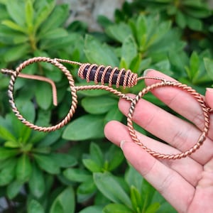 Könnte beinhalten: Eine dekorative Brille aus gedrehtem Kupferdraht mit einer zylindrischen, dunkelvioletten Steinperle, die am Steg mit Kupferdraht umwickelt ist. Die Brillengläser sind kreisförmig und durch einen einzelnen horizontalen Drahtsteg verbunden.