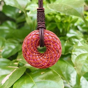 May include: A circular pendant featuring a deep red, textured stone center wrapped in a complex, spiraling pattern of thin, dark orange cord. The pendant is suspended from a thick, dark brown braided cord with a decorative knot detail.