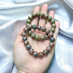 May include: Two beaded bracelets with speckled, earth-toned beads. The beads are primarily brown and green, with flecks of blue, yellow, and white. The bracelets are displayed on a light-colored surface.