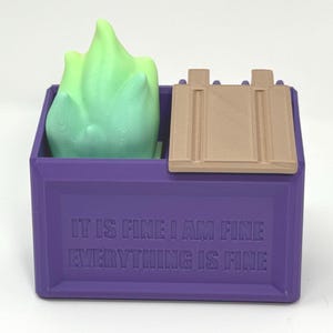 May include: A purple box with the text "IT IS FINE I AM FINE EVERYTHING IS FINE" printed on the front. Inside the box is a light green object. A tan lid is partially covering the box.