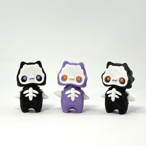 May include: Three small, stylized skeleton figures. Two are black with white and purple accents, and one is purple with white and orange accents. Each figure has a skeleton design on the torso and a cat-like head.