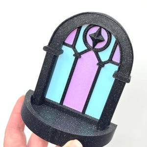 May include: A black, arched window-shaped decorative item with a stained-glass design. The window features a star in the center and is filled with blue and purple panels. The item has a small shelf at the bottom.