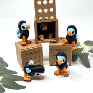 May include: A collection of 3D-printed dodo bird figurines. The birds have blue bodies, orange beaks and feet, and are wearing black hooded cloaks. They are posed around and inside small brown wooden crates.