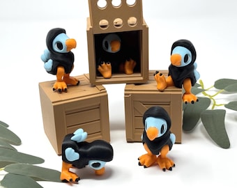 Op de afbeelding: Een verzameling 3D-geprinte dodo vogelbeeldjes. De vogels hebben blauwe lichamen, oranje snavels en poten en dragen zwarte capuchons. Ze zijn geplaatst rond en in kleine bruine houten kisten.