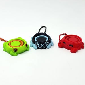 May include: Three colorful, small, round containers with attached keychains. One is green with orange and yellow accents, another is black and blue, and the third is red. Each has a unique design and a small, circular opening.
