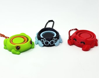 May include: Three colorful, small, round containers with attached keychains. One is green with orange and yellow accents, another is black and blue, and the third is red. Each has a unique design and a small, circular opening.