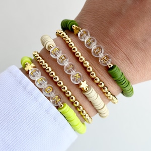 May include: A set of five bracelets with gold beads and various charms. The bracelets are stacked on a person's wrist. The charms include a star, a clover, and letters. The bracelets are made from different materials, including clear beads, green beads, and white beads.