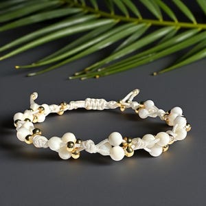 May include: White beaded bracelet with gold accents. The bracelet features a macrame design with white beads and small gold-colored beads. The bracelet is on a dark gray surface with a green leaf in the background.