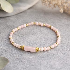 Peut inclure: Un bracelet de perles roses délicat avec des détails dorés. Le bracelet est composé de petites perles roses rondes et d'éléments dorés. Une pierre rose rectangulaire est centrée, encadrée de détails dorés ornés. Un bijou élégant.
