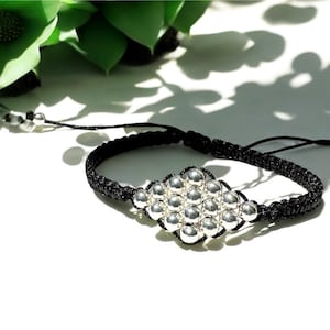Könnte beinhalten: Schwarzes Makramee-Armband mit einem silbernen rautenförmigen Mittelstück. Das Armband hat ein gewebtes Design und verstellbare Gleitknoten. Das silberne Mittelstück besteht aus mehreren kleinen, runden Perlen. Der Hintergrund ist weiß mit grünem Laub.