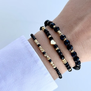 May include: Three black beaded bracelets with gold accents. The middle bracelet features a small gold heart charm.