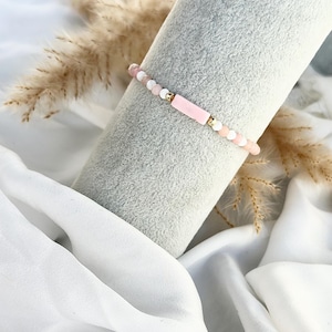 Pode incluir: Uma pulseira delicada com uma combinação de contas rosa, brancas e douradas. A pulseira tem uma peça central retangular rosa e é exibida em um objeto cilíndrico cinza com um fundo de tecido branco. É uma joia.