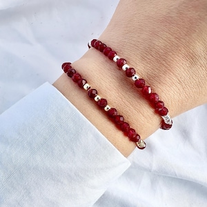 Op de afbeelding: Twee armbanden met dieprode edelstenen en zilveren en gouden accenten. De armbanden worden om een pols gedragen, waarbij de ene armband iets boven de andere zit. De achtergrond is een wit overhemd.