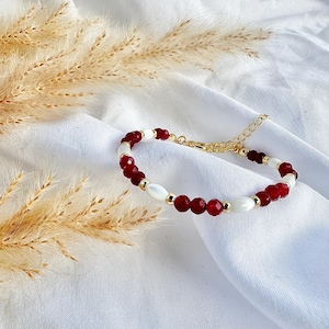Könnte beinhalten: Ein goldfarbenes Armband mit abwechselnd roten und weißen Perlen. Das Armband hat eine zarte Kette und einen Verschluss. Die Perlen sind eine Mischung aus runden und ovalen Formen. Das Armband wird auf weißem Stoff präsentiert.