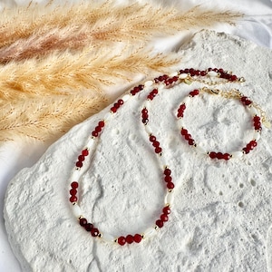 May include: A necklace and bracelet set featuring red and white beads with gold accents. The jewelry is displayed on a textured white surface, with dried ornamental grass in the background. The necklace and bracelet are connected with gold clasps.