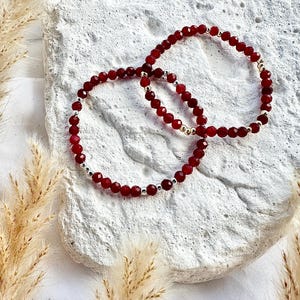 Puede incluir: Dos pulseras de cuentas rojas con detalles plateados se muestran sobre una superficie blanca texturizada. Las pulseras están hechas de pequeñas cuentas rojas facetadas ensartadas con cuentas plateadas. Son una joya.