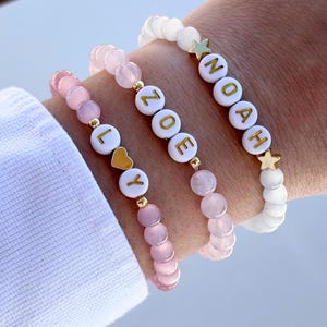 Könnte beinhalten: Drei Perlenarmbänder mit den Namen "Lily", "Zoe" und "Noah", die in goldenen Buchstaben geschrieben sind. Das "Lily"-Armband hat einen Herzanhänger, das "Zoe"-Armband hat rosa Perlen und das "Noah"-Armband hat weiße Perlen und Sternanhänger.