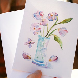 Op de afbeelding: Een aquarel schilderij van een boeket roze en paarse tulpen in een blauwe vaas. Een vlinder vliegt in de buurt van de vaas. Het schilderij is op een witte achtergrond.